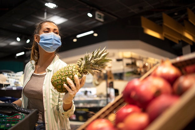 코로나 바이러스 기간 동안 과일을 구입하고 유행성 격리를 준비하는 식료품 점에서 위생 마스크와 고무 장갑 및 장바구니를 가진 여성