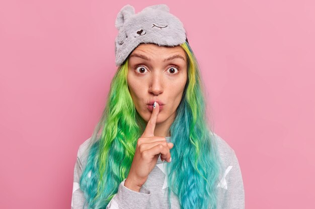  woman with long dyed hair makes silence or taboo gesture tells secret information wears sleepmask and pajama poses on pink 