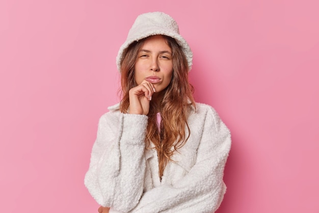 woman with long hair keeps lips folded hand on chin looks mysteriously at camera wears fur white hat and coat isolated on pink. Clothes concept