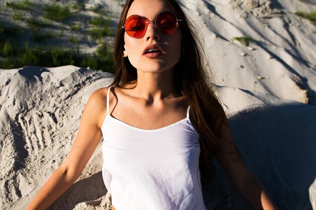 Woman with long hair in red sunglasses sits on white sand