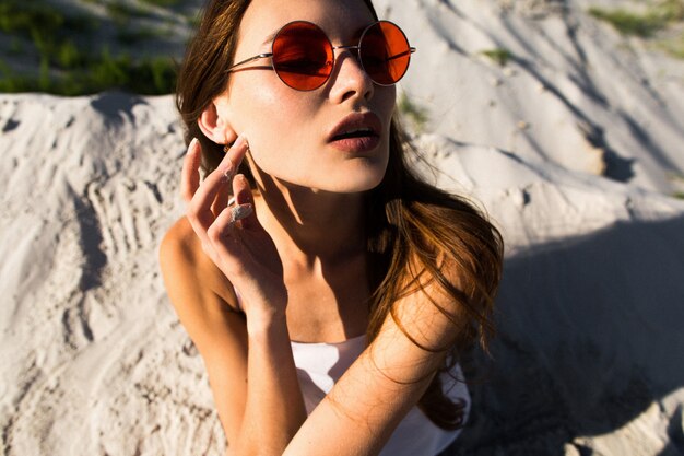 Женщина с длинными волосами в красных солнцезащитных очках сидит на белом песке