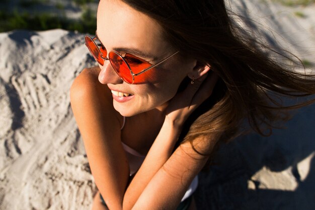 Woman with long hair in red sunglasses sits on white sand