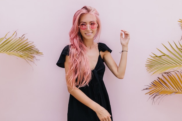 Free photo woman with long pink hair standing beside exotic palm trees in studio.