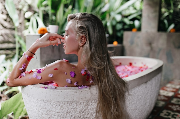 woman with long straight hair sitting in bath full of rose petals. Indoor shot of magnificent tanned woman resting at home and doing spa.