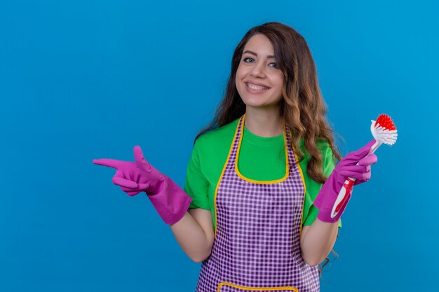 woman with long wavy hair in apron and gloves holding scrubbing brush pointing to the side smiling cheerfully standing on blue