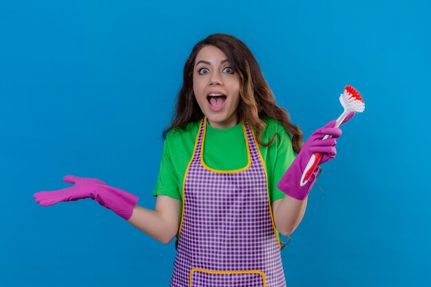 женщина с длинными волнистыми волосами в фартуке и перчатках держит щетку для мытья посуды, стоя с широко открытыми глазами и ртом, выглядя возбужденной и удивленной на синем