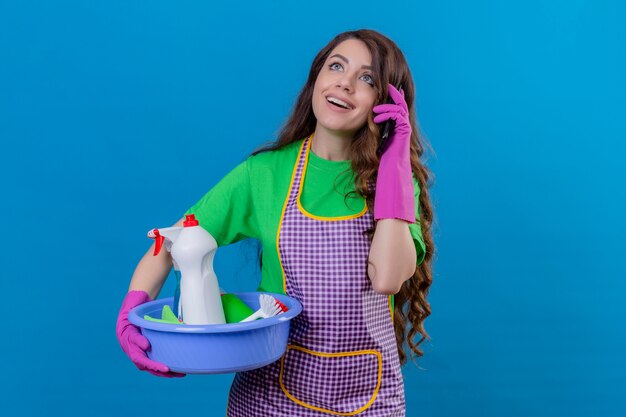 ブルーに立っている幸せそうな顔に笑みを浮かべて携帯電話で話しているツールを洗浄する洗面器を保持しているエプロンとゴム手袋を着用して長いウェーブのかかった髪を持つ女性