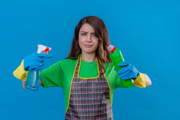 Free photo woman with long wavy hair wearing apron and rubber gloves holding bottles of cleaning supplies in raised hands looking uncertain having doubt trying to make choice standing on blue