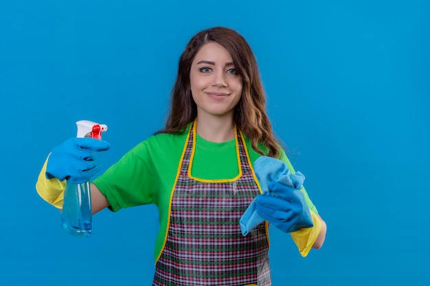 женщина с длинными волнистыми волосами в фартуке и резиновых перчатках держит коврик и чистящий спрей, выглядит уверенно и позитивно улыбается, готовая к уборке, стоя на синем