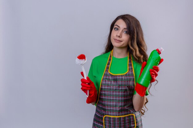 Donna con lunghi capelli ondulati che indossa un grembiule e guanti di gomma tenendo la spazzola per strofinare e una bottiglia con prodotti per la pulizia con sorriso scettico in piedi