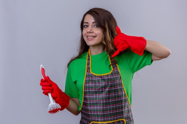 Donna con lunghi capelli ondulati che indossa grembiule e guanti di gomma che tiene spazzola per strofinare facendo chiamarmi gesto guardando fiducioso sorridente amichevole