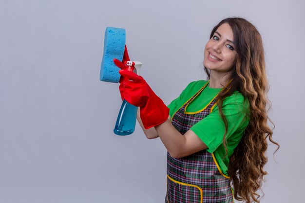 woman with long wavy hair wearing apron and rubber gloves holding sponge and cleaning spray ready ready for cleaning smiling with happy face standing