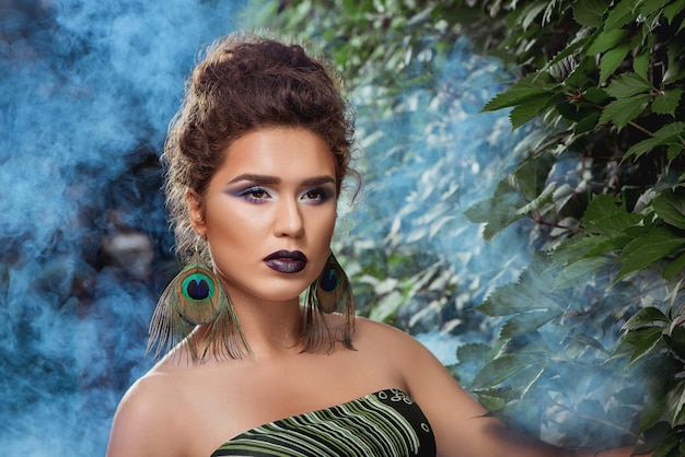 Free photo woman with make up posing against green bush in blue smoke