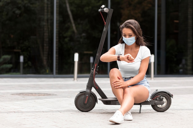 電動スクーターに座ってスマートウォッチをチェックする医療マスクを持つ女性