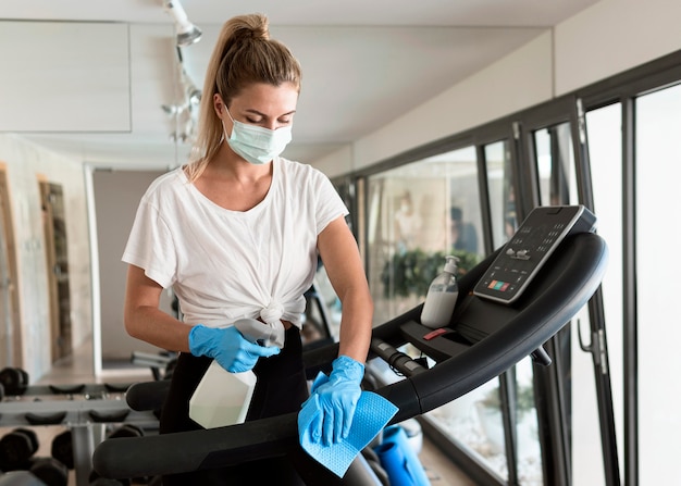 Woman with medical mask and cleaning solution disinfecting gym equipment