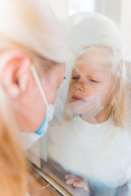 Free photo woman with medical mask in quarantine behind window with cute girl