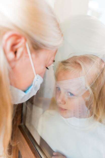 Woman with medical mask in quarantine behind window with little girl