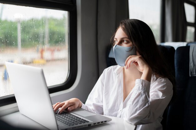Woman with medical mask traveling by public train and using laptop