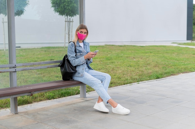 Foto gratuita donna con maschera medica che utilizza smartphone mentre aspetta l'autobus pubblico