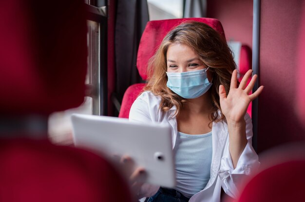 Женщина в медицинской маске с помощью планшета для видеозвонка во время поездки на общественном поезде