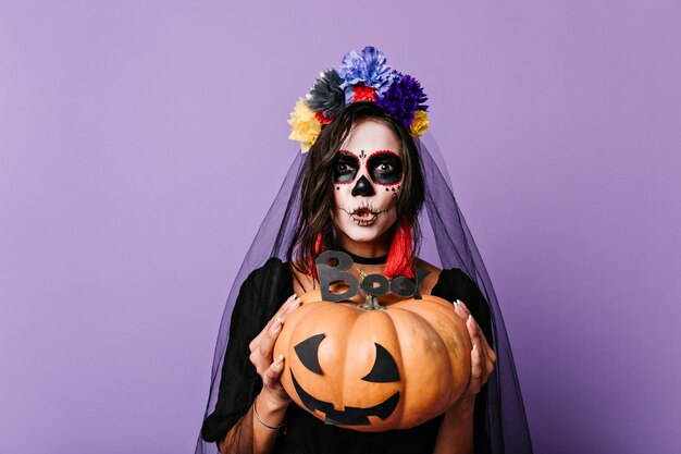 Woman with Mexican-style face art is trying to scare. Brunette with pumpkin and black wedding veil posing on lilac wall.