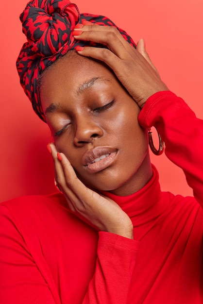 woman with natural beauty without makeup keeps eyes closed touches gently face wears kerchief tied on head and turtleneck isolated on vivid red