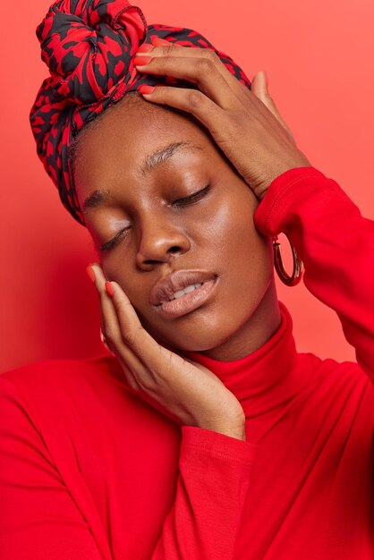 woman with natural beauty without makeup keeps eyes closed touches gently face wears kerchief tied on head and turtleneck isolated on vivid red