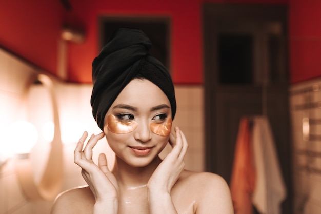 Woman with patches under eyes and towel on her head posing against wall of bathroom