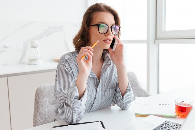 woman with pensive look holding pencil and talking on smartphone while siting at workplace in white room