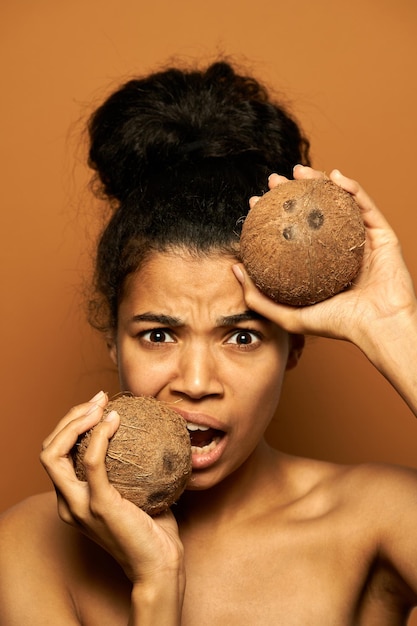 woman with perfect skin and hair in a bun looking at camera, posing with two coconuts near her face isolated on orange