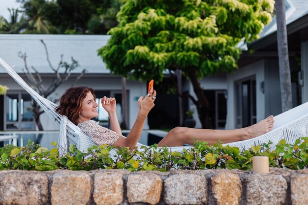 Woman with phone resting lying on hammock with mobile phone