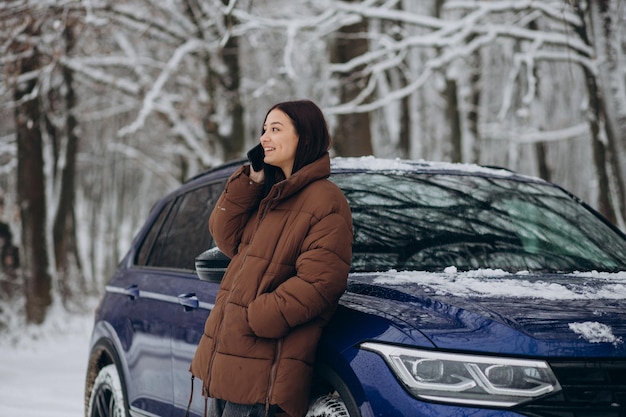 冬​の​森​で​彼女​の​車​の​そば​に​立っている​電話​を​持つ​女性