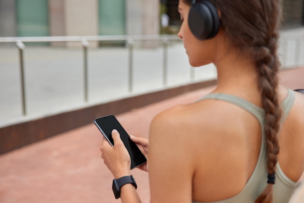 woman with pigtail uses mobile phone checks fitness activity chooses song to listen for workout wears wireless headphones on ears dressed in active wear stands outdoors