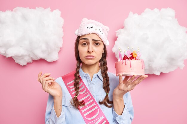 woman with pigtails looks worried wears sleepmask shirt and birthday ribbon poses indoor isolated on pink