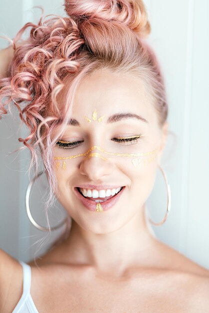 ピンクの髪とペイントストロークの形で芸術的なメイクアップの女性が笑顔で目を閉じ、明るい白でポーズをとる
