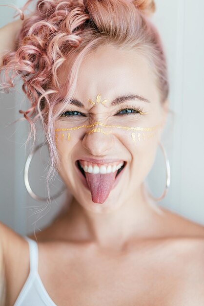 woman with pink hair and artistic makeup smiling at camera, sticking her tongue out, posing on light white. Face art concept