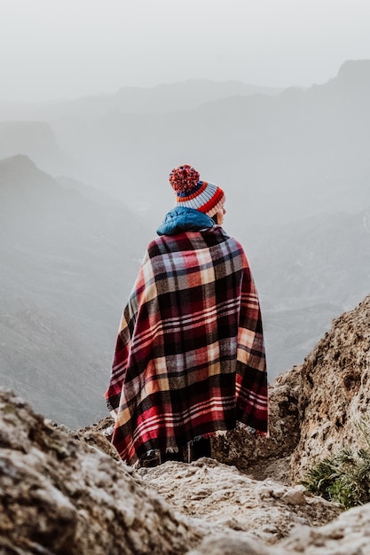 Donna con poncho scozzese e berretto con pompon che si affaccia su uno splendido paesaggio di montagna