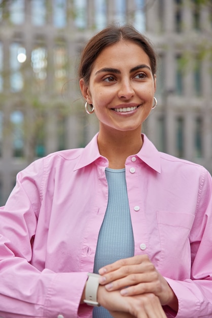 женщина с приятной внешностью ждет, когда кто-то назначит встречу, проверяет время на часах, улыбается, нежно стоит на улице на размытом архитектурном здании, носит розовую рубашку.