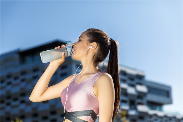 Woman with a ponytail is drinking from the sportive bottle in the earphones near the modern building