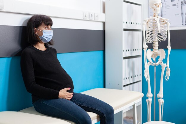 キャビネットでの診察予約時にフェイスマスクを着用し、医師の診察を待っている妊娠中の腹を持つ女性。健康管理検査のためにオフィスに座っている赤ちゃんを期待している患者 Premium写真