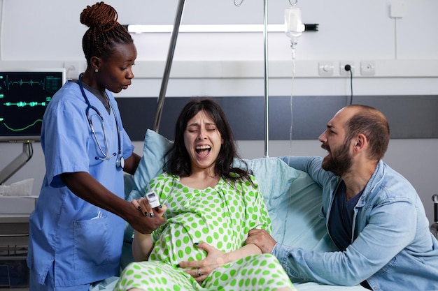 Woman with pregnancy pain getting medical assistance from african american nurse in hospital ward. Future father standing beside pregnant wife supporting and comforting her about childbirth