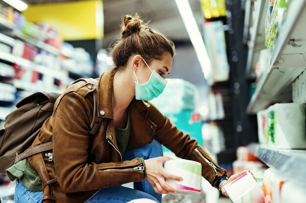 바이러스 전염병 동안 슈퍼마켓에서 화장지를 쇼핑하는 보호용 안면 마스크를 쓴 여성