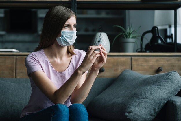 Woman with protective mask sitting on sofa holding syringe