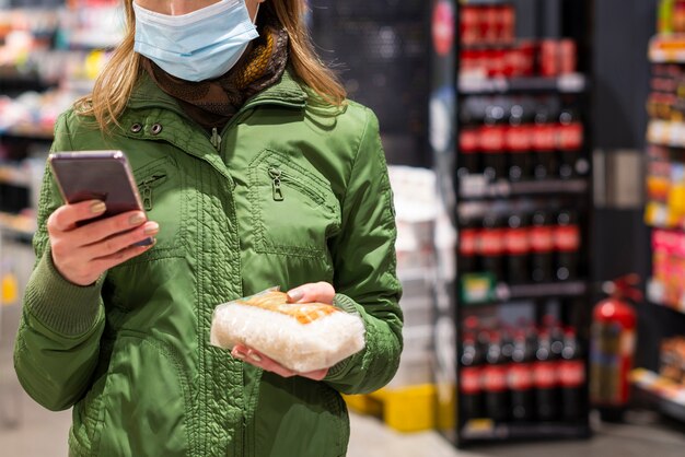 Женщина с защитной маской, используя свой мобильный телефон в магазине