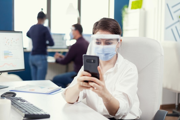 Женщина с защитной маской, работающая в профессиональной рабочей области, набирая текст на мобильном телефоне перед компьютером