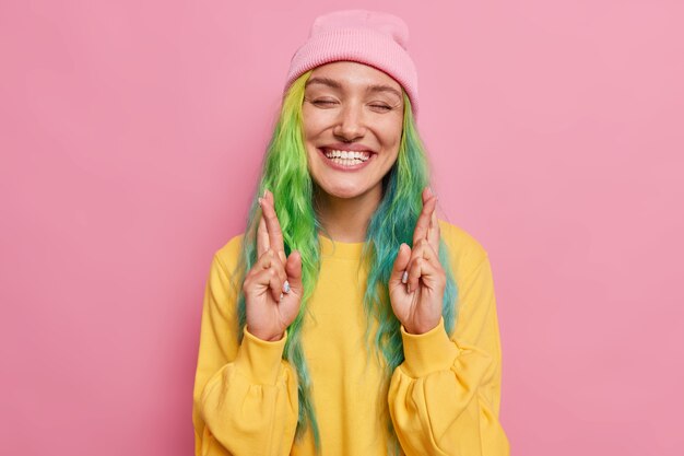 虹のヘアスタイルを持つ女性は目を閉じて指を交差させます幸運の笑顔を信じて気持ちよく素晴らしい結果を期待していますピンクの壁に隔離された帽子とジャンパーを着ています