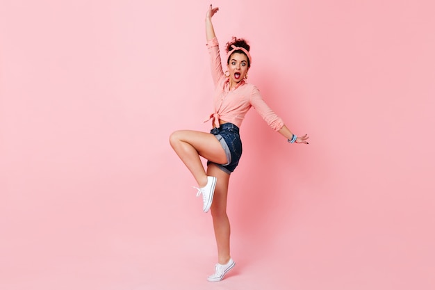 Woman with red lips and blue eyes in surprise looks at camera, raises her leg and hands on pink space. Portrait of pin-up lady.