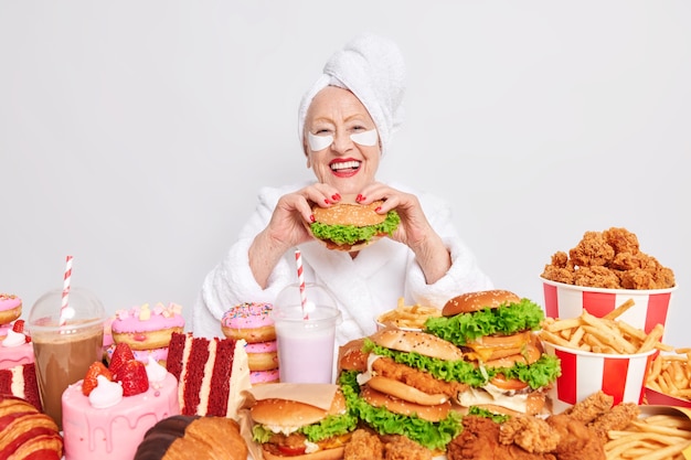 La donna con le labbra rosse si diverte a mangiare un gustoso hamburger dipendente dal cibo spazzatura non segue la dieta applica cerotti di bellezza sotto gli occhi pose a tavola ha fame