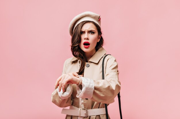 Woman with red lips and green eyes looks indignantly at camera. Portrait of girl wearing felt hat and trench.