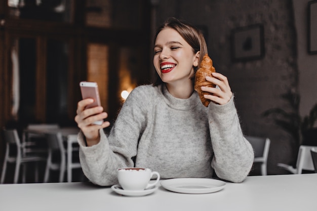 Woman with red lipstick is laughing while chatting in smartphone. Portrait of lady in gray sweater with croissant in her hands.